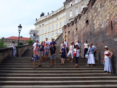 Chór, w letnich, niezobowiązujących strojach przechadza się po starych uliczkach Pragi. Zdjęcie zrobione jest na tzw. schodach zamkowych. Zrobione jest z dołu, chórzyści stoją na schodach kilka stopni wyżej, w luźnej, rozsypanej szeroko grupie. W tle, wysoko, widoczny jest zespół zamkowy Hradczany.