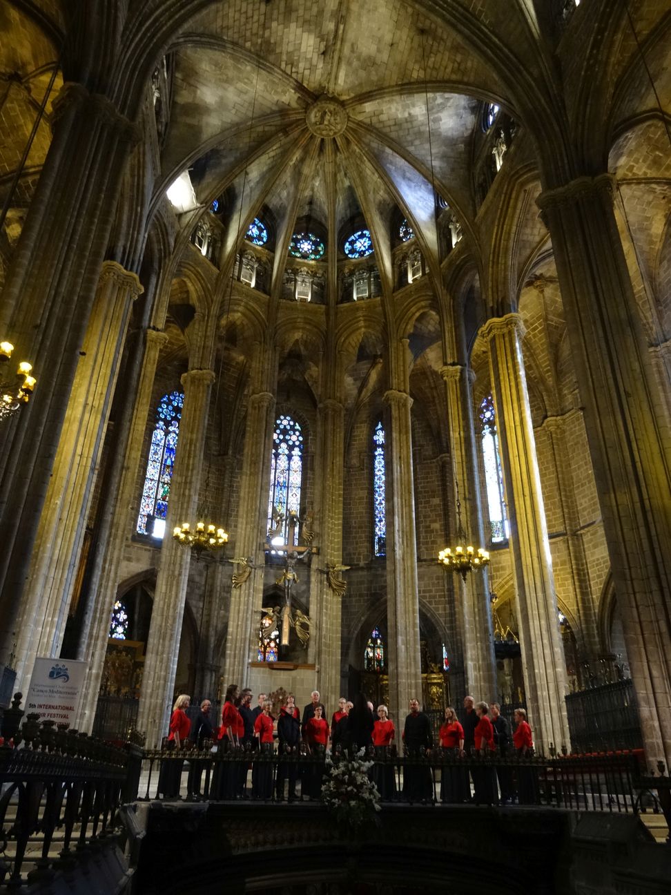 Bardzo światowe zdjęcie. Stoimy na nim w prezbiterium gotyckiej Katedry św. Eulalii w Barcelonie, która jest ogromna. Zdjęcie zrobione jest nieco od dołu, tak, że widać nie tylko nas ale i całą, ogromną wysokość prezbiterium łącznie z kawałkiem gotyckiego sklepienia. Widoczne są kolumny, wysokie gotyckie okna z witrażami, świecące żyrandole i krzyż nad tabernakulum. Jesteśmy na tym tle bardzo mali. Stoimy za kutymi, czarnymi balustradami, które oddzielają prezbiterium od reszty kościoła. Stoimy w półkolu, przemieszani. Tyłem do fotografa stoi Ewa, która dyryguje. Chórzystki mają na sobie czerwone bluzki i grafitowe spódnice, panowie są w czarnych koszulach z czerwonymi muchami. W kościele panuje półmrok.