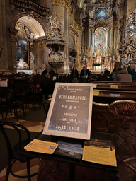 Plakat informujący o naszym koncercie, umieszczony na specjalnym stojaku wewnątrz starego, barokowego kościoła na starym mieście w Wiedniu. Tytuł koncertu to "Veni Emmanuel". W tle rozświetlony ołtarz, kilka osób siedzących w kościelnych ławkach i przechodzący turyści, zwiedzający świątynię.