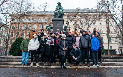 Beethoven siedzi w dostojnej pozie na cokole. W tle budynek najstarszego gimnazjum w Wiedniu i hotel Ritz. Chór stoi przed pomnikiem na trzech stopniach podestu.