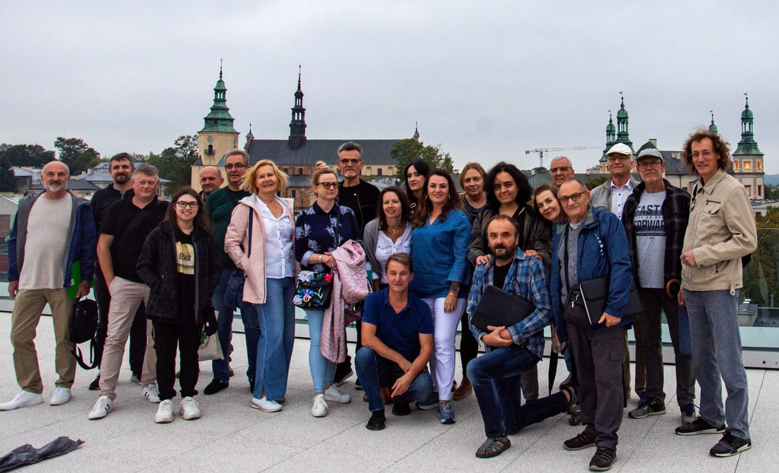 Chórzyści stoją na rozległym tarasie znajdującym się na dachu Teatru Żeromskiego. Barierki tego tarasu są szklane, więc za nimi widoczne jest miasto - Wzgórze Katedralne i Pałac Biskupów Krakowskich.