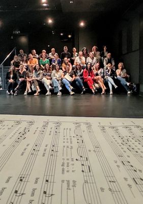 Na pierwszym planie są nuty, chórzyści są za nimi w oddali. Siedzą w sali prób Teatru Żeromskiego, na szerokich schodach, które się w tej sali znajdują.