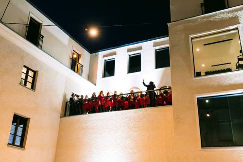Chórzyści stoją wysoko na tarasie, sfotografowani są z dołu, z pozycji widza stojącego na dziedzińcu. Zapada już wieczór więc niebo jest czarne, a taras, ściany teatru i śpiewający chórzyści są jasno oświetleni reflektorami.