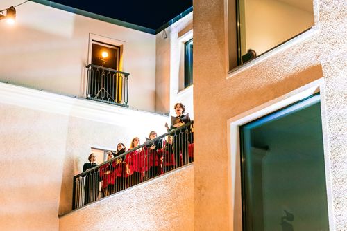 Chórzyści stoją wysoko na tarasie, sfotografowani są z dołu, z pozycji widza stojącego na dziedzińcu. Zapada już wieczór więc niebo jest czarne, a taras i ściany teatru są jasno oświetlone reflektorami, w świetle których bardzo jaskrawo wyróżniają się czerwone tuniki chórzystek. Na pierwszym planie Marek Saramak, student Katedry Muzyki UJK, który gościnnie towarzyszy nam jako solista.